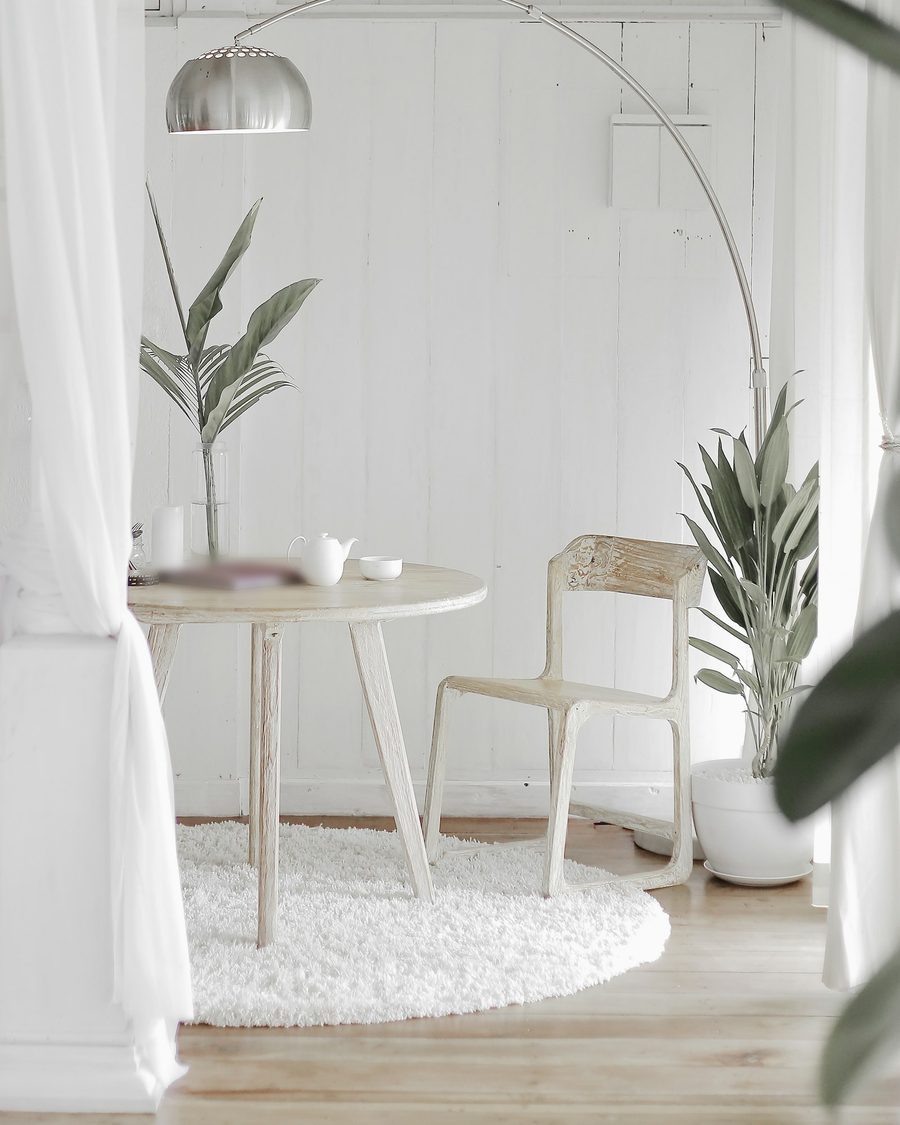 Heller Raum mit weißen Holzwänden, rundem Holztisch, einem Stuhl, Bogenlampe und zwei Pflanzen